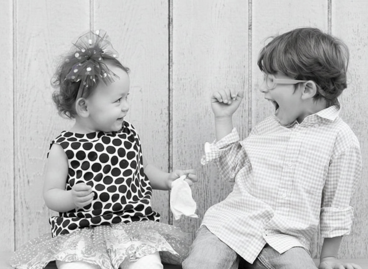 Two children sitting on a bench, one in a polka dot dress with a purple bow and the other in a checkered shirt and jeans.